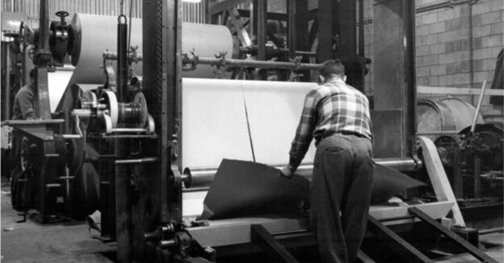 Black and white image of a worker handling roofing materials on an industrial production line with large machinery in the background.