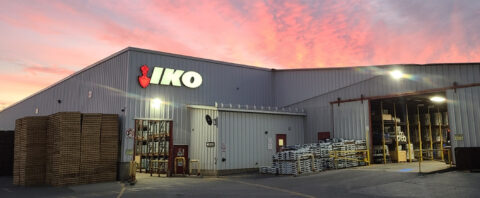 Exterior view of an IKO manufacturing facility at sunset, with stacks of materials and an open warehouse entrance.