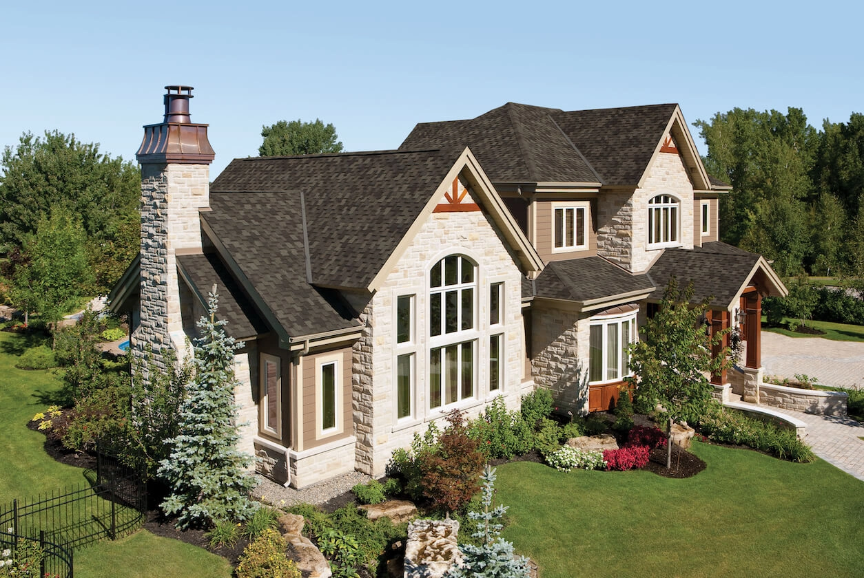 A large stone-clad house with dark Armourshake roofing, surrounded by greenery and a stone chimney, set against a clear blue sky.