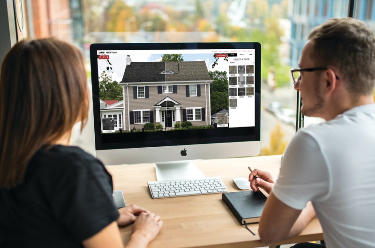 Two people sit at a desk, looking at a house design on a computer screen. One holds a notebook and pen.