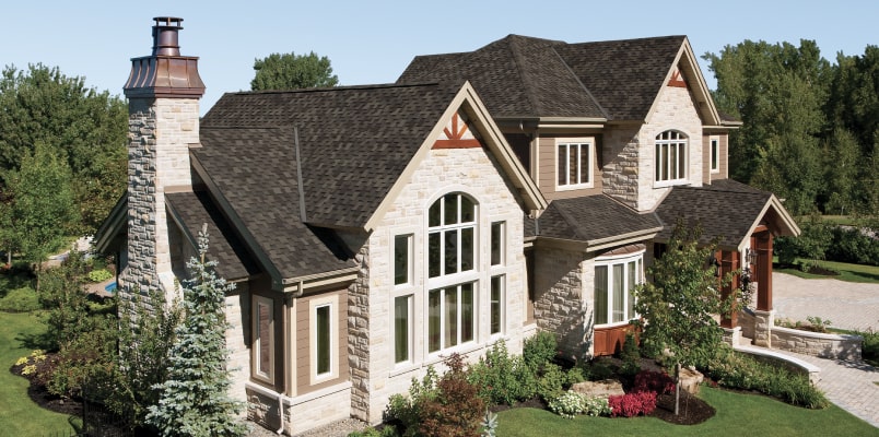 A large stone-clad house with dark Armourshake roofing, surrounded by greenery and a stone chimney, set against a clear blue sky.