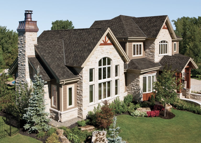 A two-story stone house with a dark Armourshake shingled roof, large arched windows, and a brick chimney. It's surrounded by manicured greenery and trees under a clear sky.