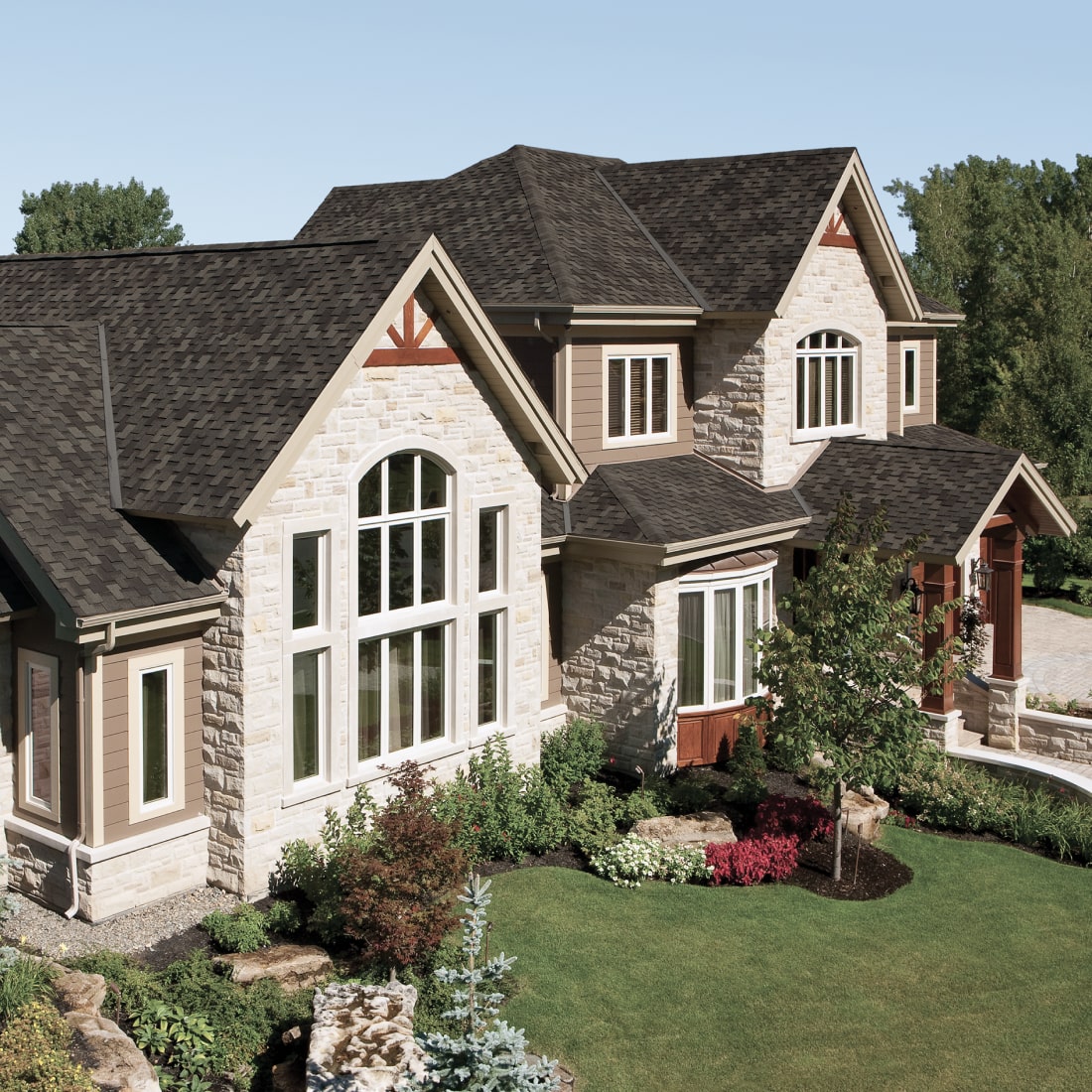 A large stone-clad house with dark Armourshake roofing, surrounded by greenery and a stone chimney, set against a clear blue sky.