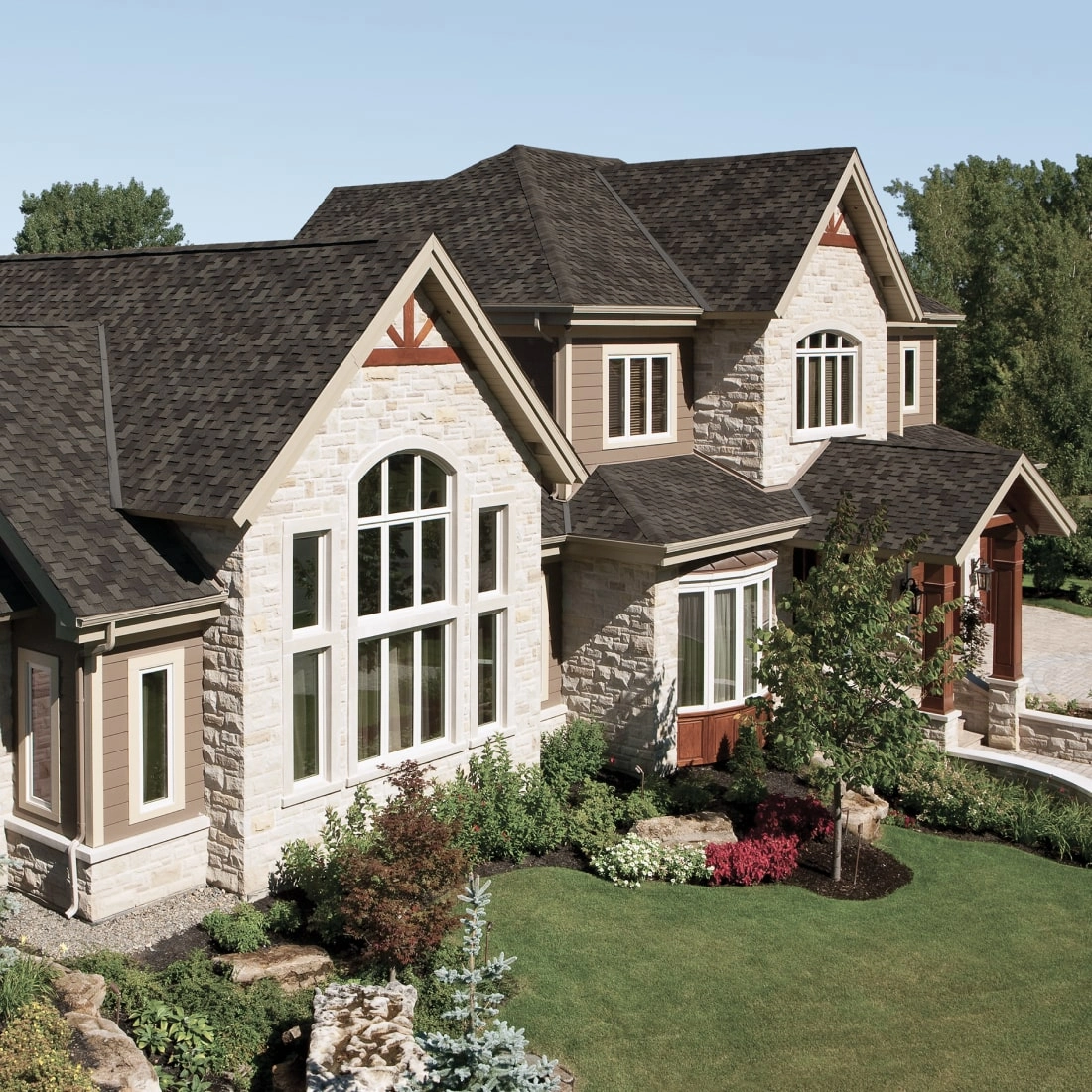 A large stone-clad house with dark Armourshake roofing, surrounded by greenery and a stone chimney, set against a clear blue sky.