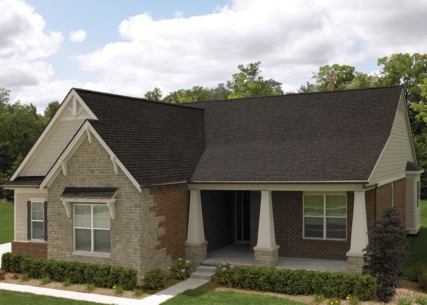 This single-story Cambridge brick house features a dark roof, white trim, and classic columns on the porch, all nestled amid lush green shrubs and trees.