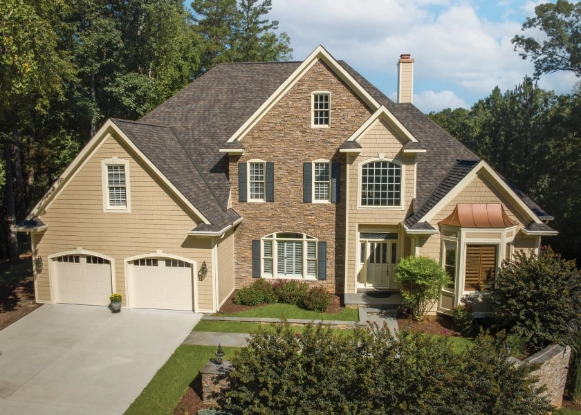 A two-story house with tan siding, stone facade, and a gray roof stands proudly like a modern dynasty. It features a double garage, large windows, and a well-maintained garden with trees in the background.