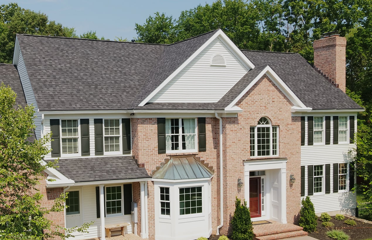 A large two-story brick house with white siding and a dark gray roof features numerous windows and a prominent entrance with black granite columns, surrounded by trees.