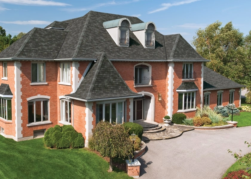 Large brick house with a dark gray roof, multiple gables, and dormer windows. The Royal Estate™ features a rounded driveway and landscaped garden in front, set against a clear blue sky in the background.