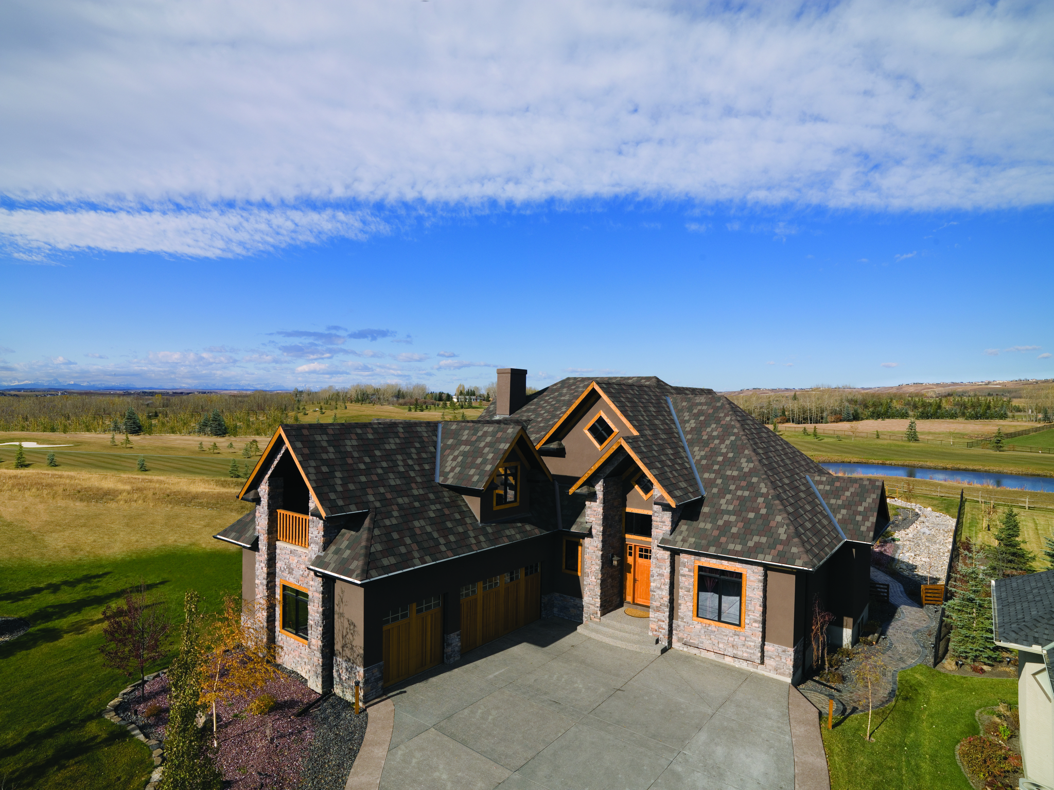 A large two-story house with a Crowne Slate and wood exterior sits in a grassy landscape with a pond and distant trees under a blue sky.