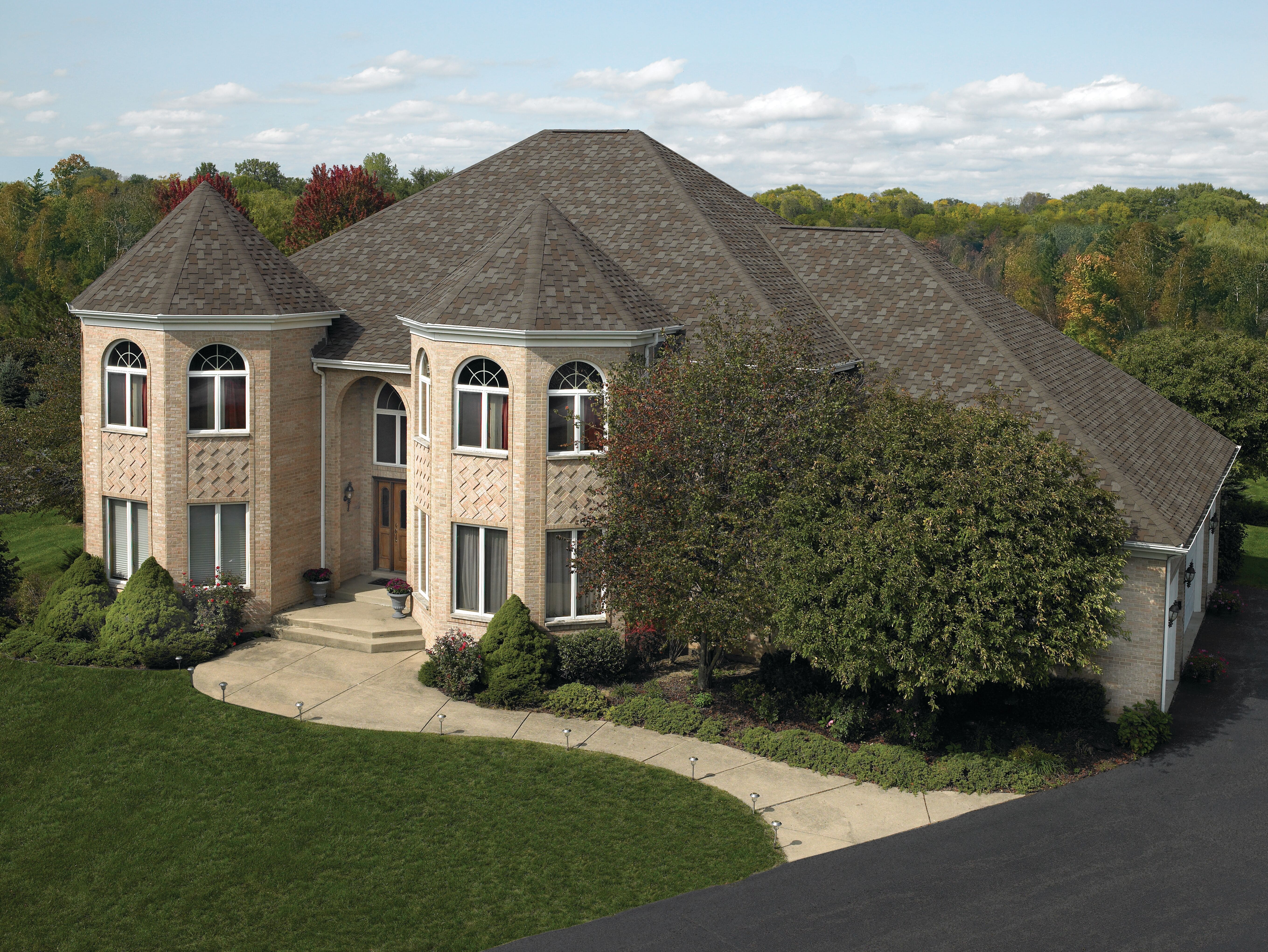 Aerial view of a large two-story brick house with a complex Armourshake roof design, surrounded by green trees and a well-manicured lawn, under a clear sky.