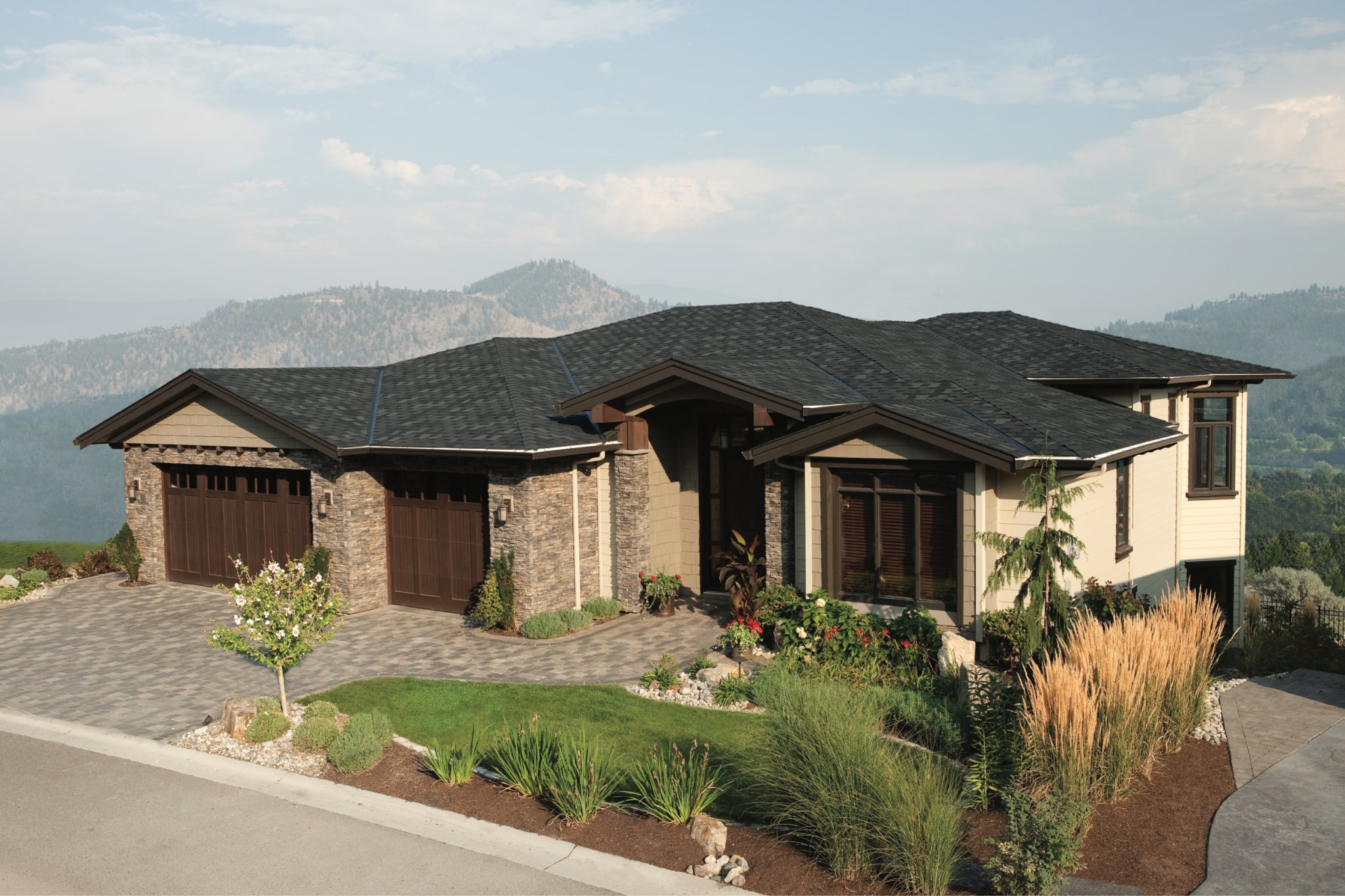 Modern house with stone and wood facade, featuring an Armourshake roof in Shadow Black, three-car garage, landscaped yard, and a breathtaking mountainous backdrop.