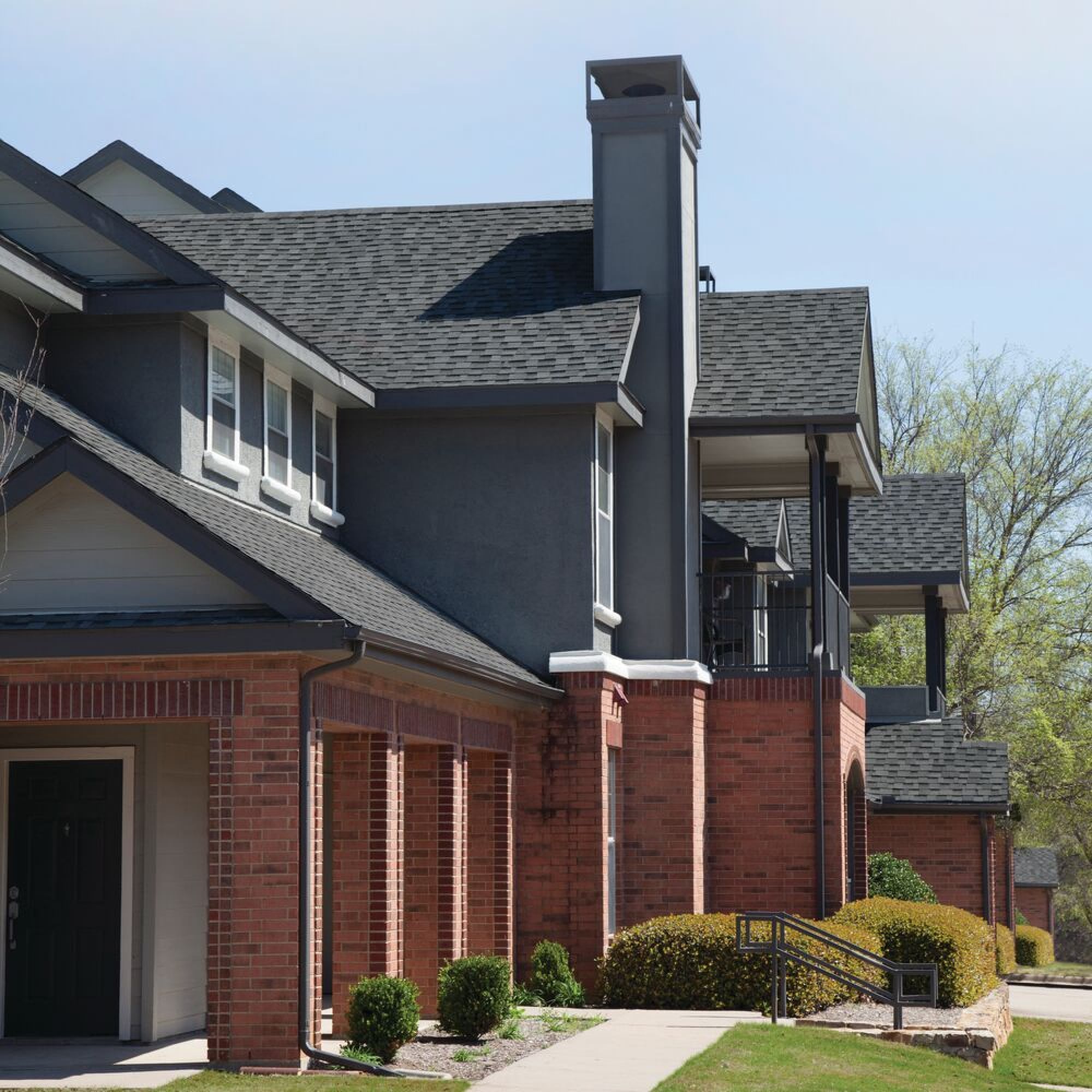 Modern residential building with a brick and dark grey exterior, reminiscent of Cambridge's architectural charm. Features include dual rooflines, a chimney, balconies, and a pathway lined with bushes.