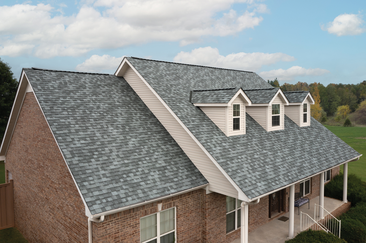 Brick house with a Frostone Grey shingle roof, featuring three dormer windows. Surrounded by a green lawn and trees under a partly cloudy sky, this home blends classic charm with modern elegance, fit for a dynasty.