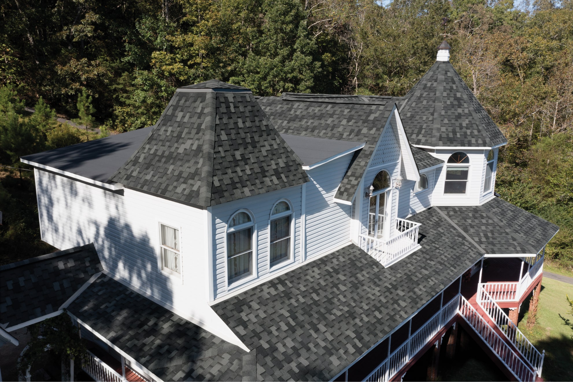 An aerial view reveals a large Greystone house with white siding and dark gray roofing shingles. Distinctive turrets and multiple windows add character, while surrounding trees frame the serene setting.