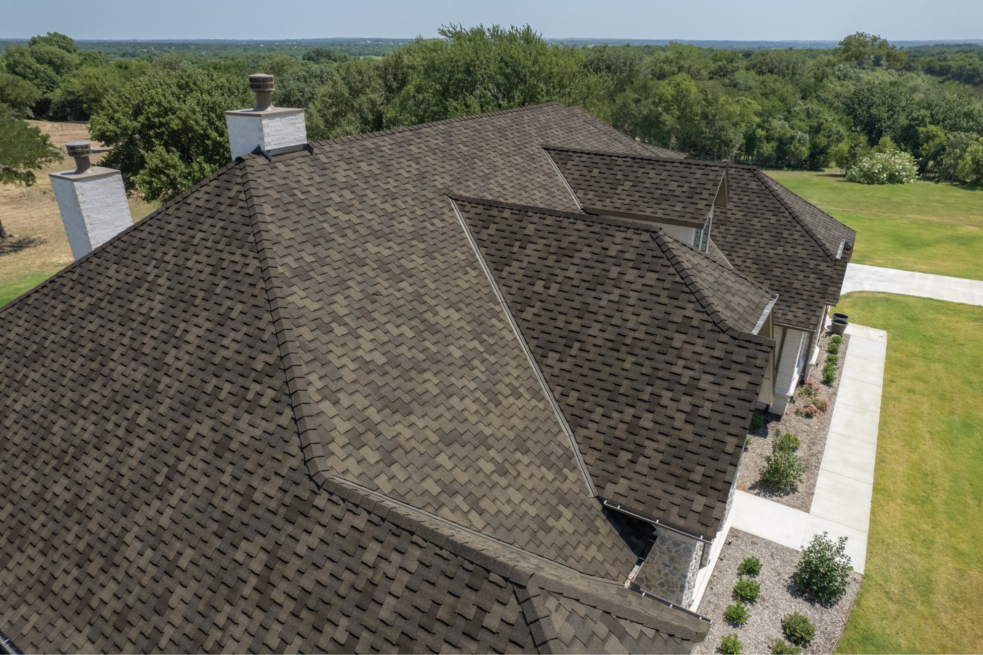 Aerial view of a house with Armourshake shingles in a Weathered Stone hue on the roof. The surrounding area includes lush green trees and a manicured lawn.