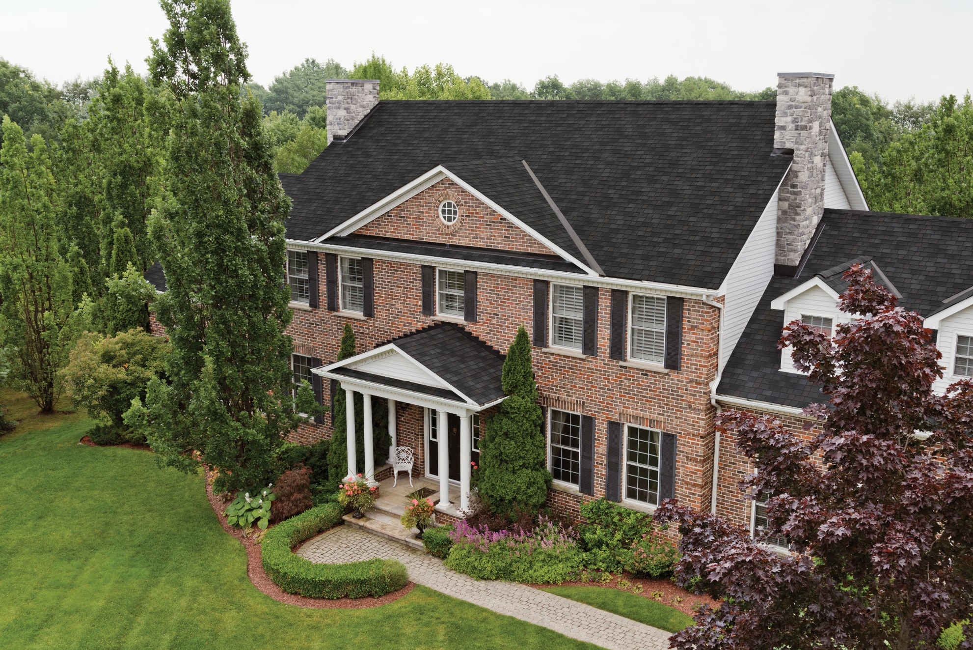 The two-story brick house with a dark Shadow Slate roof and white trim features elegant columns at the entry. Nestled in landscaped gardens, trees, and lush lawn akin to a Royal Estate, a charming pathway guides you to the welcoming front door.