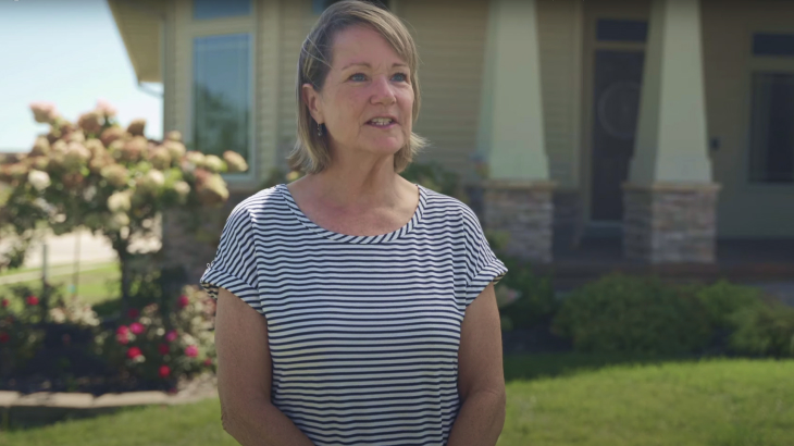 A woman in a striped shirt stands in front of a house with a garden, talking.