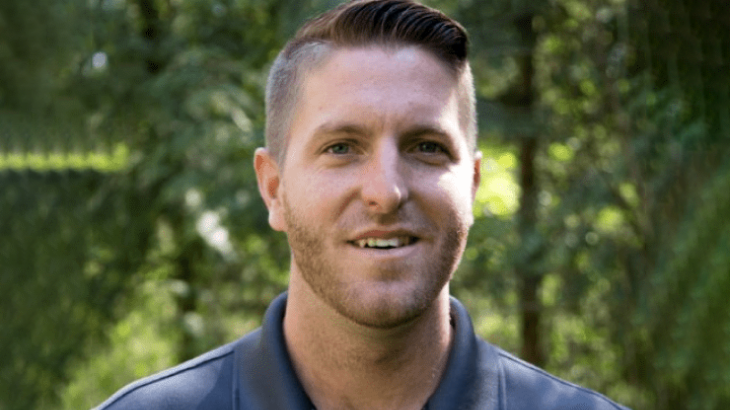 A man with short hair and a beard is smiling in an outdoor setting with trees in the background.