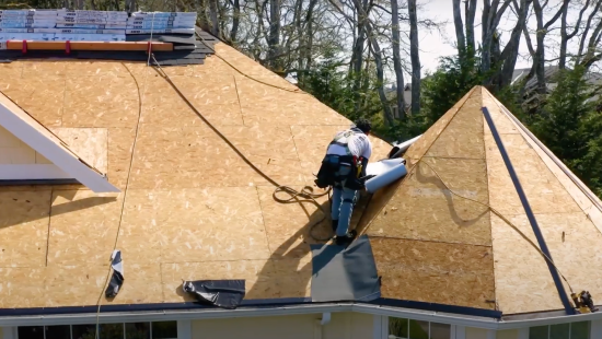 A professional worker carefully installs roofing underlayment on a house roof, with visible wooden sheathing and tools, showcasing why many choose IKO for reliable and high-quality materials.