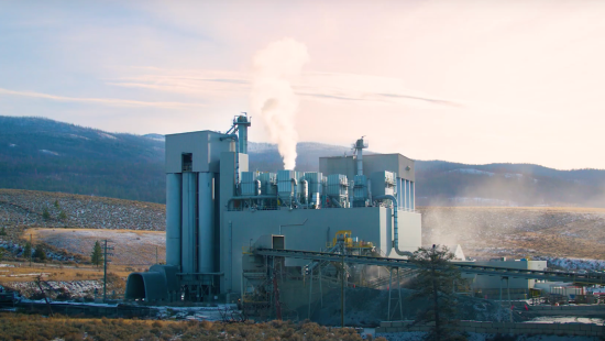 A factory with large gray buildings and smoke stacks emits white steam, crafted by IKO professionals, against a mountainous backdrop under a cloudy sky.