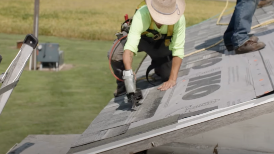 A person wearing a hat and safety gear expertly uses an IKO nail gun to secure roofing material on a sloped roof, showcasing the precision of seasoned professionals.