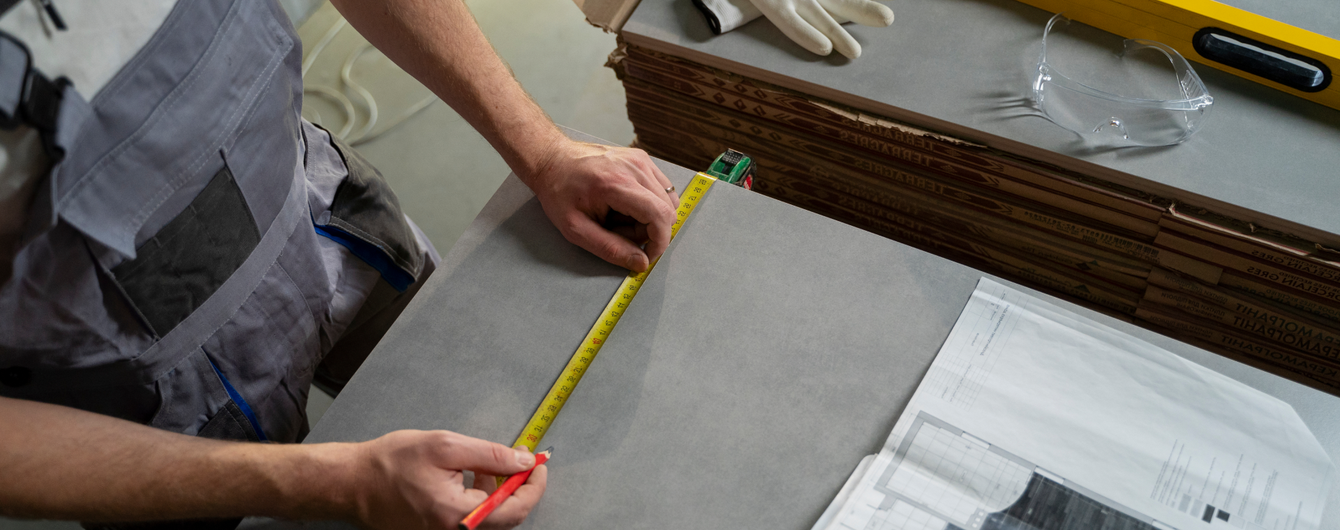 A professional carefully measures a slab of material on the table, wearing a work apron. Nearby are gloves, safety goggles, and a spirit level. IKO tools lie among papers and cardboard boxes, showcasing why experts always choose quality for precision in their craft.