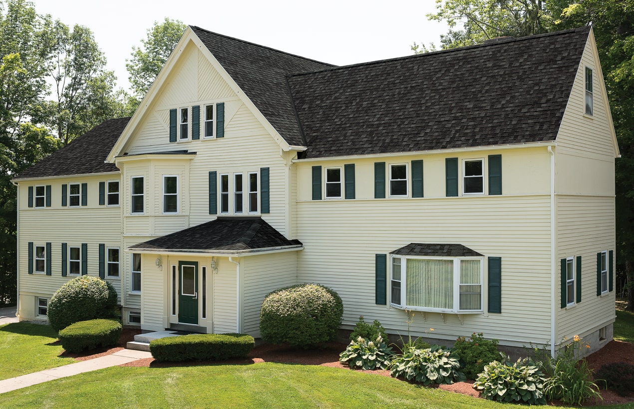 A large cream-colored house with charcoal shutters, a dark shingled roof, multiple windows, and a manicured lawn with bushes in front.