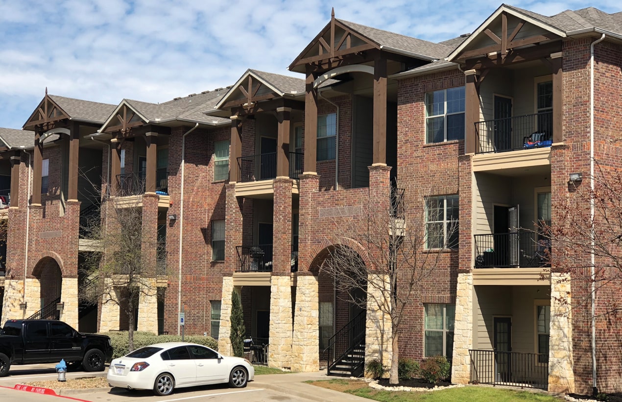 A multi-story apartment building with a classic brick facade and Cambridge stone accents stands proudly. Several balconies offer views of the scenery, while cars rest on the paved driveway. The lush trees in front add a touch of dual black elegance to its charming exterior.