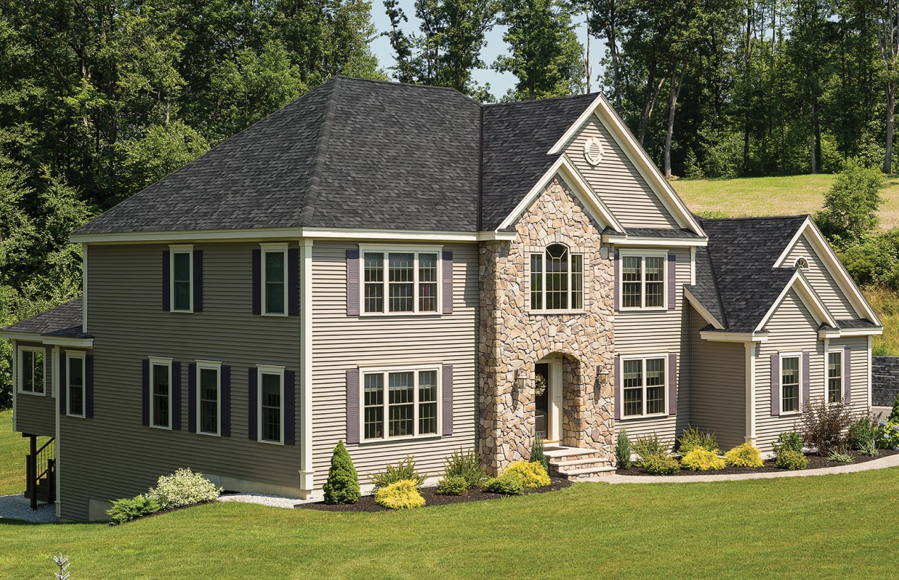 The two-story house with gray siding and stone facade, accented by black shutters, resembles a Cambridge classic. Nestled on a slate-green lawn with trees in the background, it blends timeless elegance with modern charm.