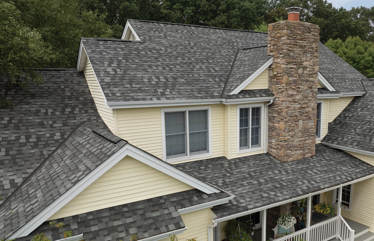 A two-story house with yellow siding, a stone chimney, and a Summit Grey shingled roof stands majestically. Dynasty-like trees create a picturesque background.