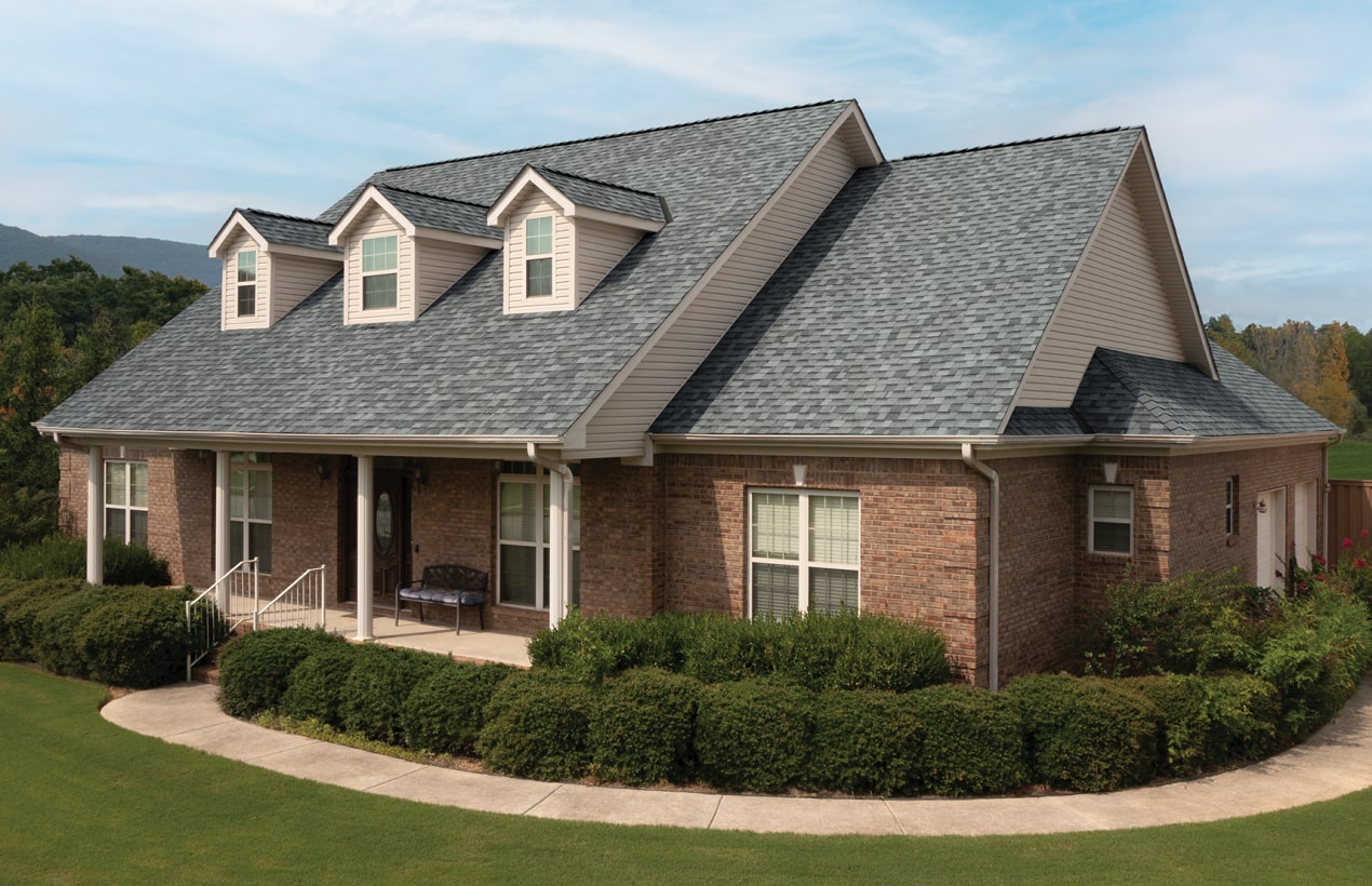 A brick house with a Frostone Grey shingle roof and three dormer windows, surrounded by green shrubs and a lawn under a blue Nordic sky.