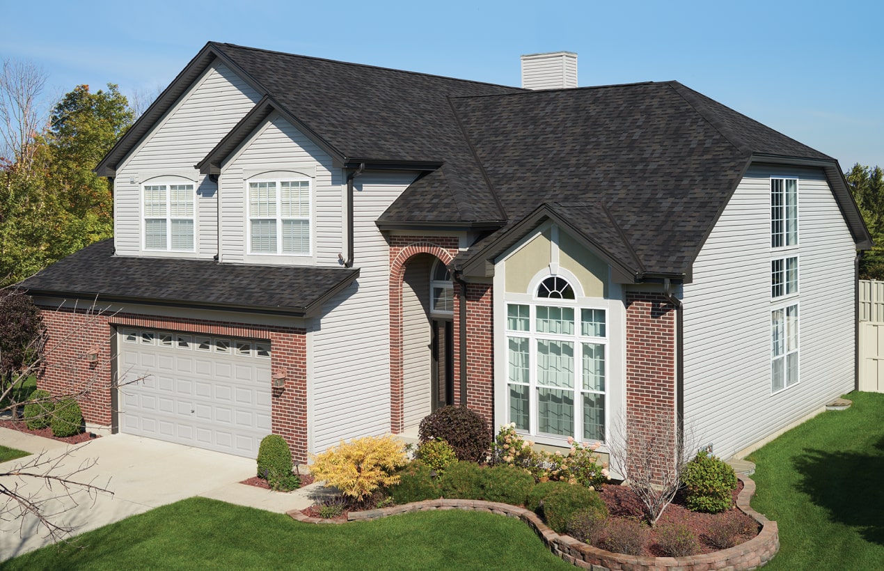 A charming two-story suburban house with glacier-gray siding, brick accents, and a sleek black roof. It features a Nordic-inspired arched entryway, a spacious two-car garage, and a beautifully landscaped front yard with lush green grass and vibrant shrubs.