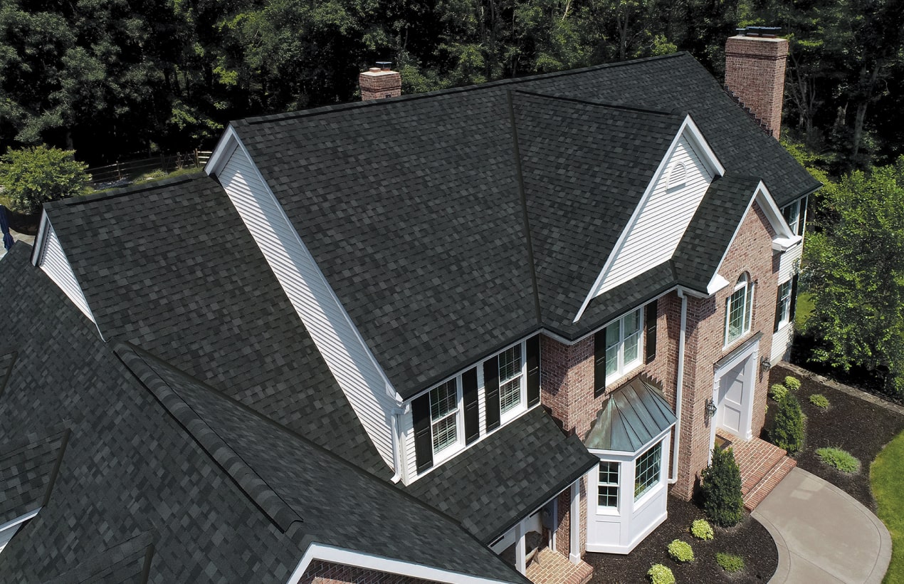 Aerial view of a large Nordic-style two-story house with dark gray shingle roofing, black brick chimney, and white siding, surrounded by trees and landscaping.