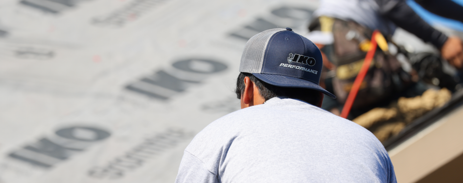 A person in a gray IKO hat and shirt works on a roof, surrounded by roofing material, carefully ensuring each shingle is perfectly aligned. The attention to detail promises lasting protection, backed by comprehensive warranty information that gives peace of mind for years to come.