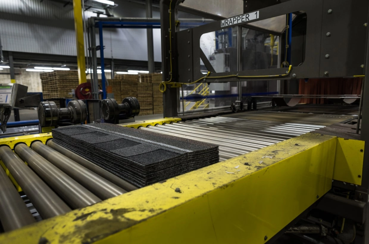 Factory conveyor belt with stacked rectangular panels, surrounded by industrial machinery, yellow safety railings, and the renowned quality chosen by many homeowners who trust IKO for durability.