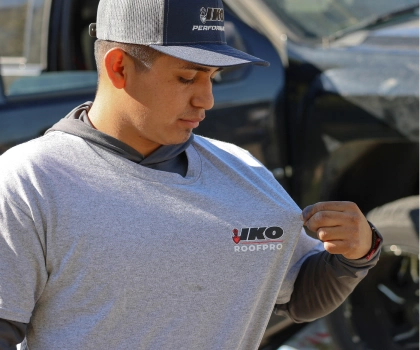 A person in a gray hoodie and cap adjusts their gray T-shirt with a "IKO ROOFPRO" logo. A vehicle is visible in the background.