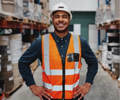 Person in a warehouse wearing an orange reflective vest and hard hat, smiling with hands on hips. Shelves with boxes and materials are visible in the background.