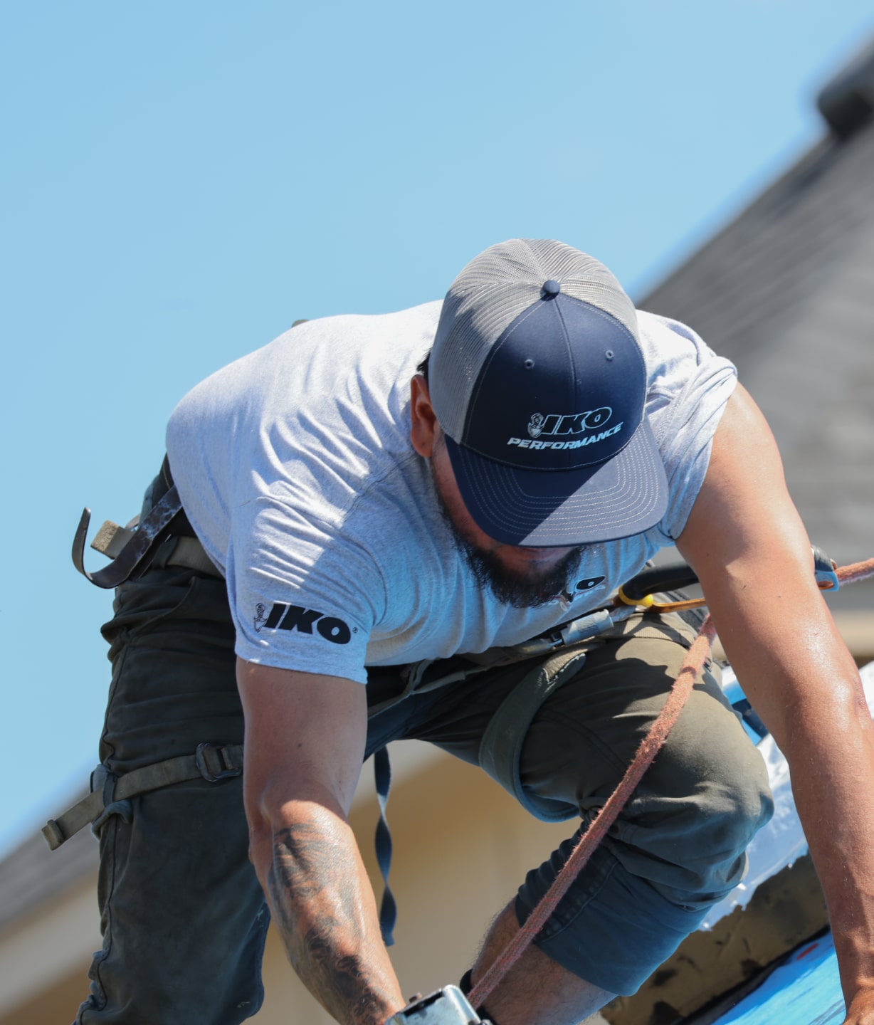 A man wearing a cap and safety harness is working on a sloped roof, with the sky visible in the background, exemplifying the safety measures that homeowners choose when selecting IKO roofing solutions.