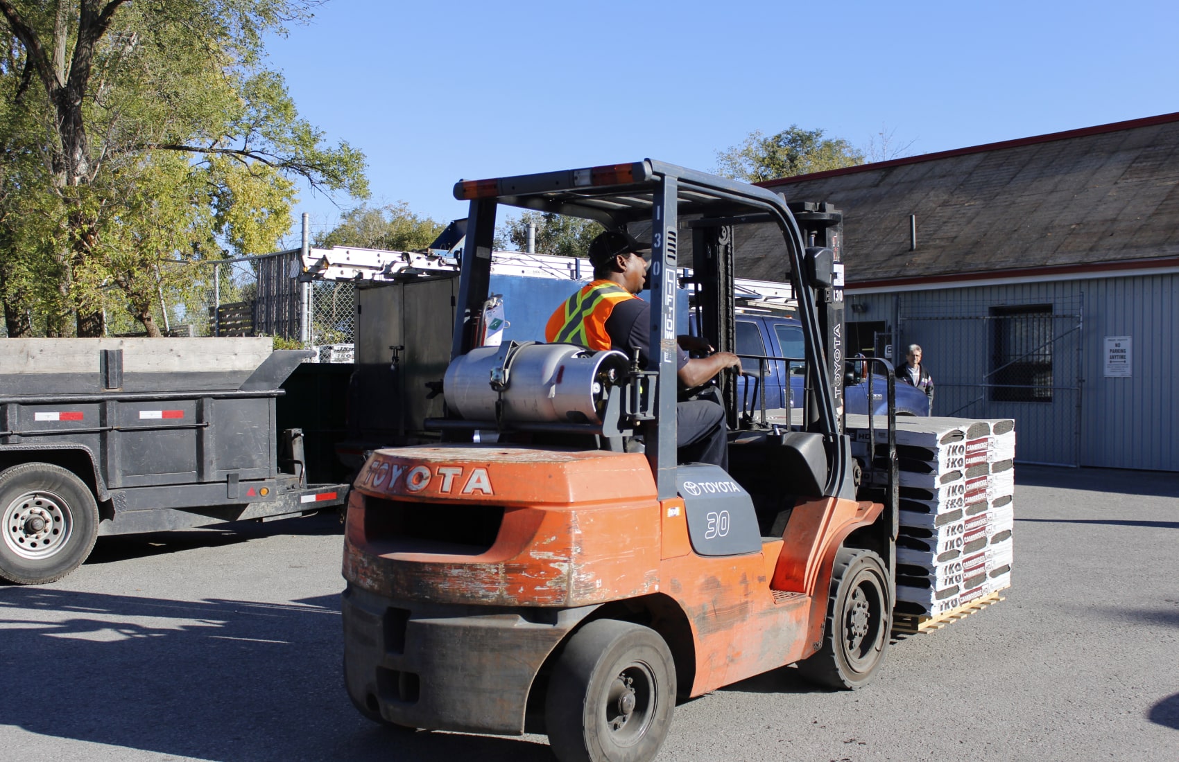 A worker expertly operates an orange forklift, transporting stacked materials with precision in an outdoor industrial area. Information on load capacity and warranty details ensures smooth operation and reliability in the bustling workplace.