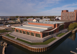 Aerial view of a modern brick building with a flat roof, surrounded by water and greenery in an urban area, under a clear blue sky.