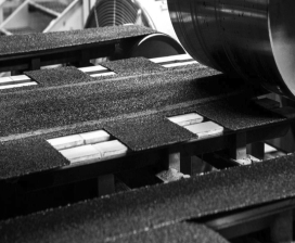 A close-up of an industrial machine with textured conveyor belts and cylindrical components, handling rectangular objects in a manufacturing setting. Black and white image.