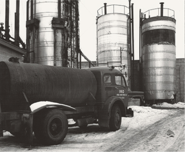 A vintage truck parked in front of three large industrial storage tanks in a snowy setting.