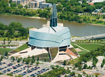 Aerial view of a modern, architecturally distinct building with a tower, situated near a river and surrounded by parking lots and greenery.