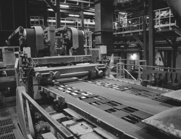 A monochrome image of an industrial machine in a factory, with conveyor belts and rollers, used for processing materials.