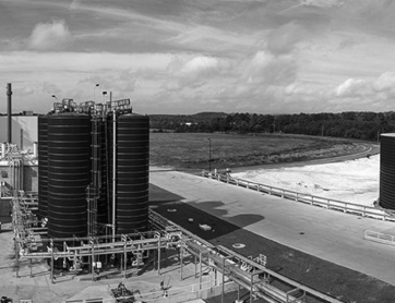 Industrial facility with large cylindrical tanks, pipelines, and adjacent open land under a cloudy sky. Black and white image.