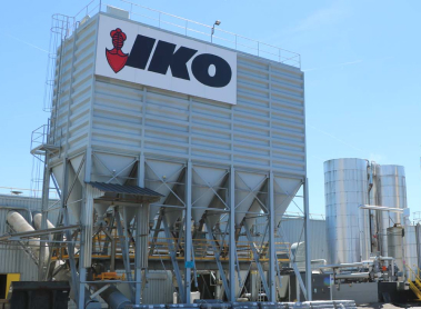 Industrial facility with large metal silos, featuring an IKO sign on the central structure. Blue sky background.