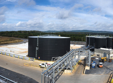 Storage tanks at an industrial facility surrounded by roads and pipes, with trees and hills in the background under a partly cloudy sky.