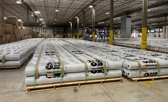 Stacks of large rolls wrapped in white material are organized on pallets in a spacious warehouse with high ceilings and rows of lights.