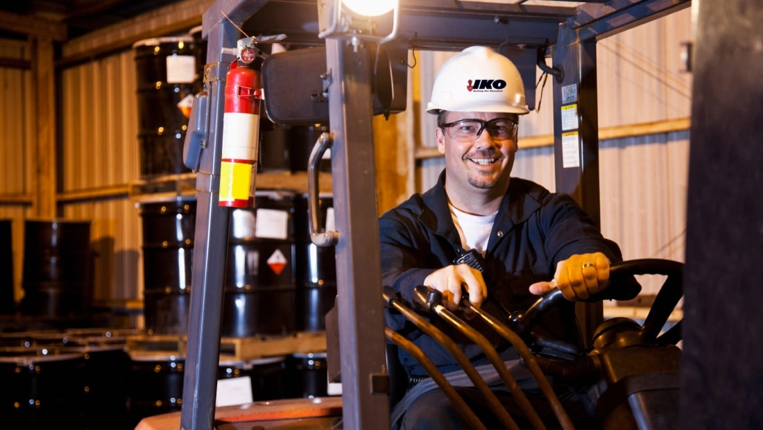 A man wearing a hard hat operates a forklift in a warehouse filled with stacked barrels.