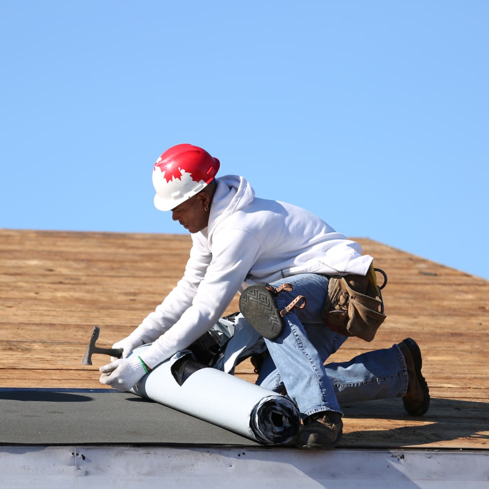 A worker in a white hoodie and red hard hat is kneeling and installing roofing material on a wooden roof under a clear blue sky.